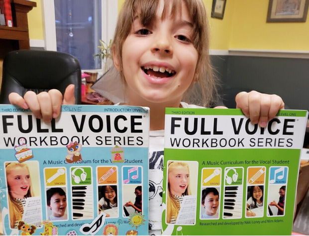 a smiling child showing her old Introductory book covered in stickers and her new Level One book.
